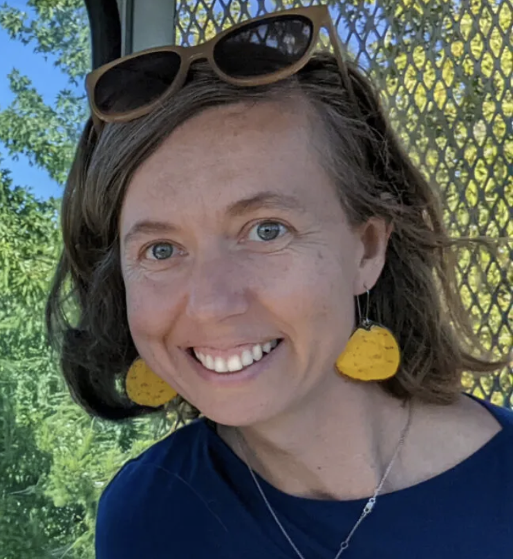 Smiling woman with shoulder-length hair, wearing yellow statement earrings, sunglasses on her head, and a navy top, standing outdoors in front of greenery and a mesh screen.